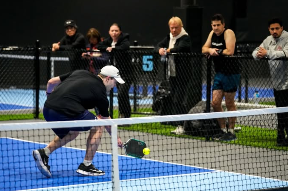Pickleball player in action reaching for a low ball with spectators behind fence